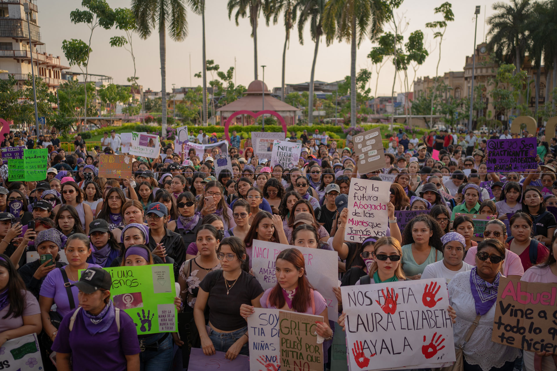 A large crowd of women gathered in a public square, holding signs with various messages. Many wear purple clothing and bandanas. Palm trees and a cityscape can be seen in the background.