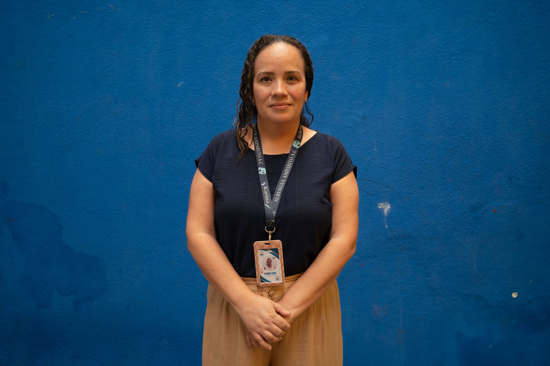 Karina del Carmen Vidal Baeza has long hair and stands against a blue wall. She is wearing a navy blue shirt and light brown pants, with an ID badge on a lanyard around their neck. Their hands are clasped in front of them, and they are looking at the camera.