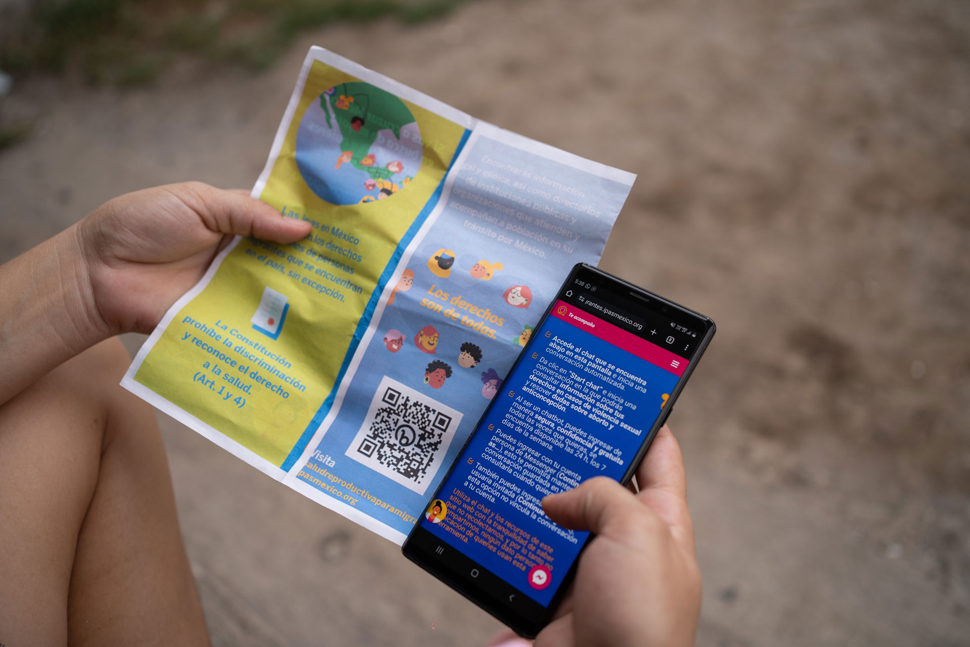 A person is holding a colorful flyer and a smartphone displaying a bright app or website. The flyer features a large QR code and various cartoon-like illustrations. The ground is visible in the background.