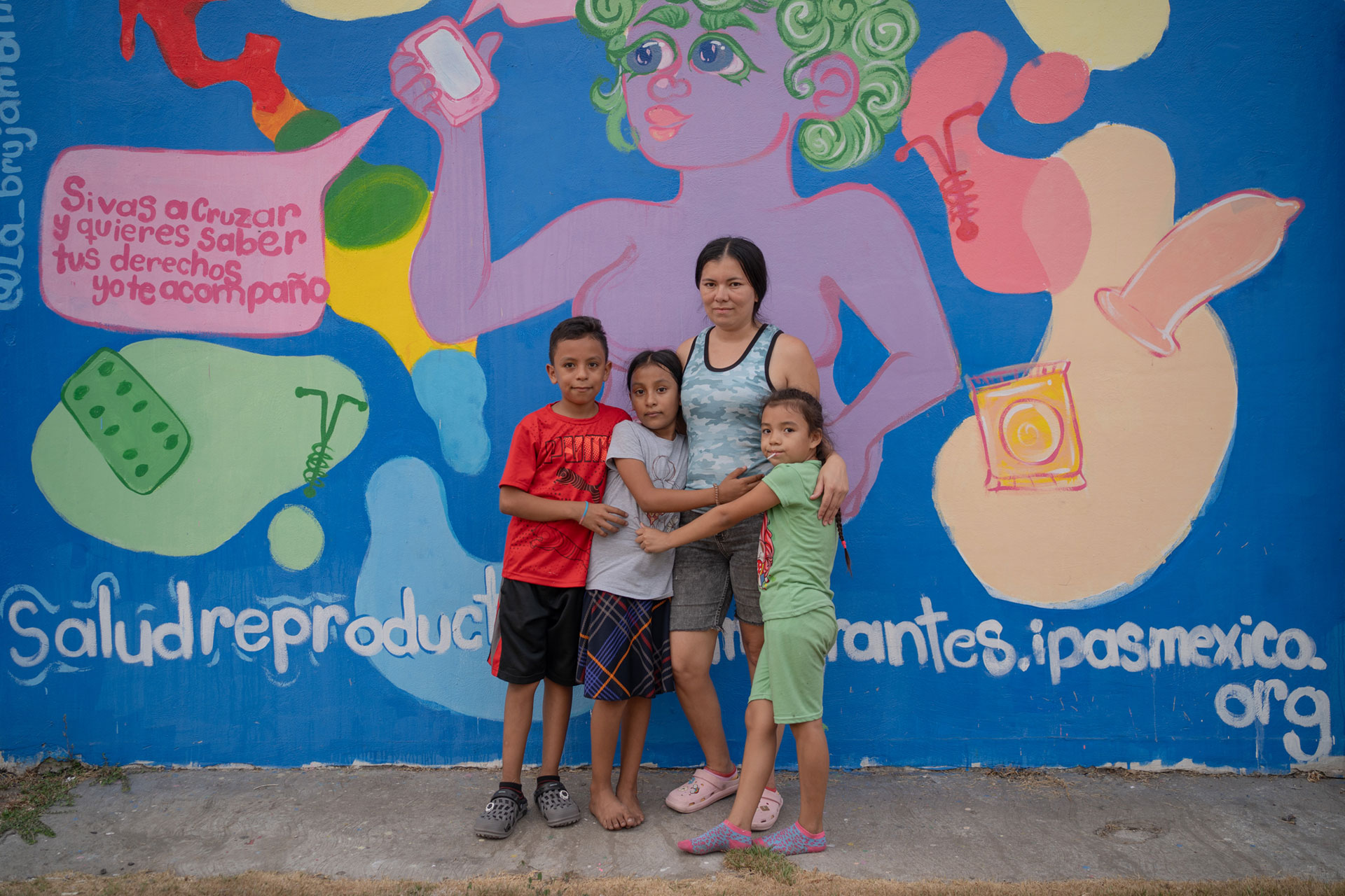 Saraí Gomez with her three children stand in front of a colorful mural featuring a large figure and text about reproductive health. The mural includes bright shapes and symbols, with the words "salud reproductiva" visible at the bottom.