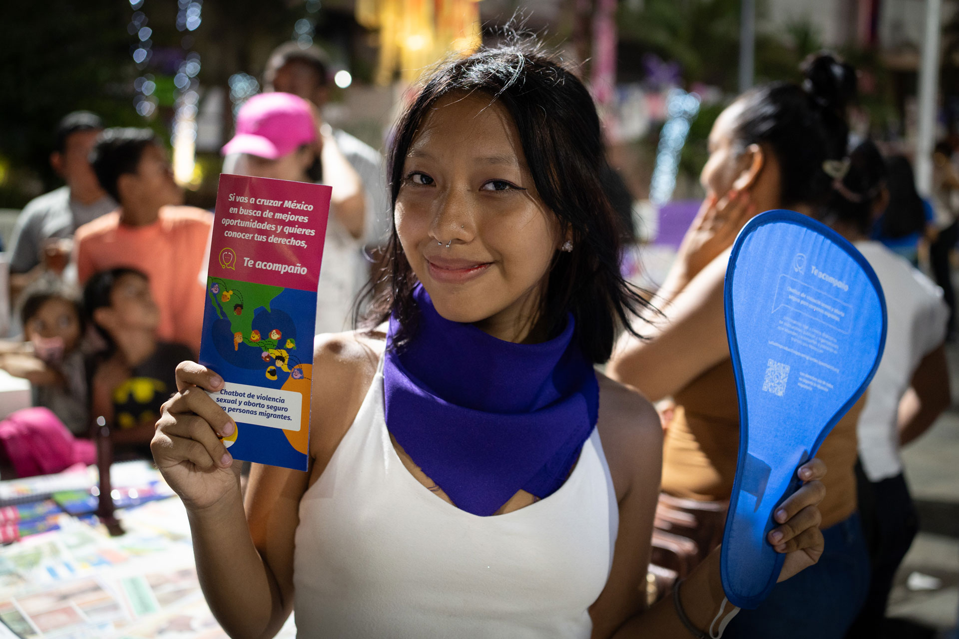 A young woman with a purple scarf smiles, holding a pamphlet and a blue fan. She stands at an event with a crowd in the background, under colorful lights.