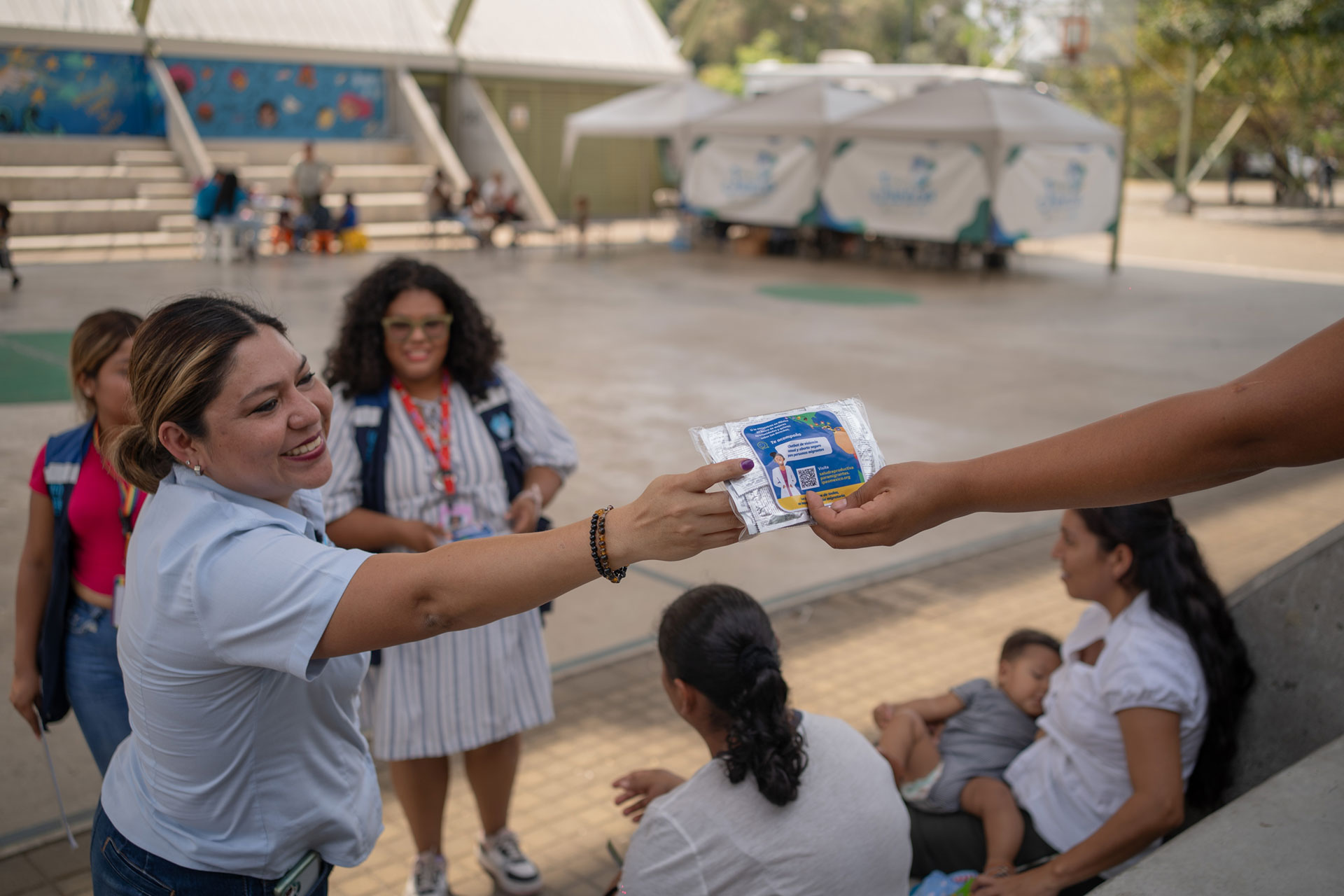 Yadira Guerrero, wearing a light blue shirt is smiling and holding out a packet to another woman on the right. They are outdoors in a park area with people sitting around, including a woman with a baby. There are structures and tents in the background.