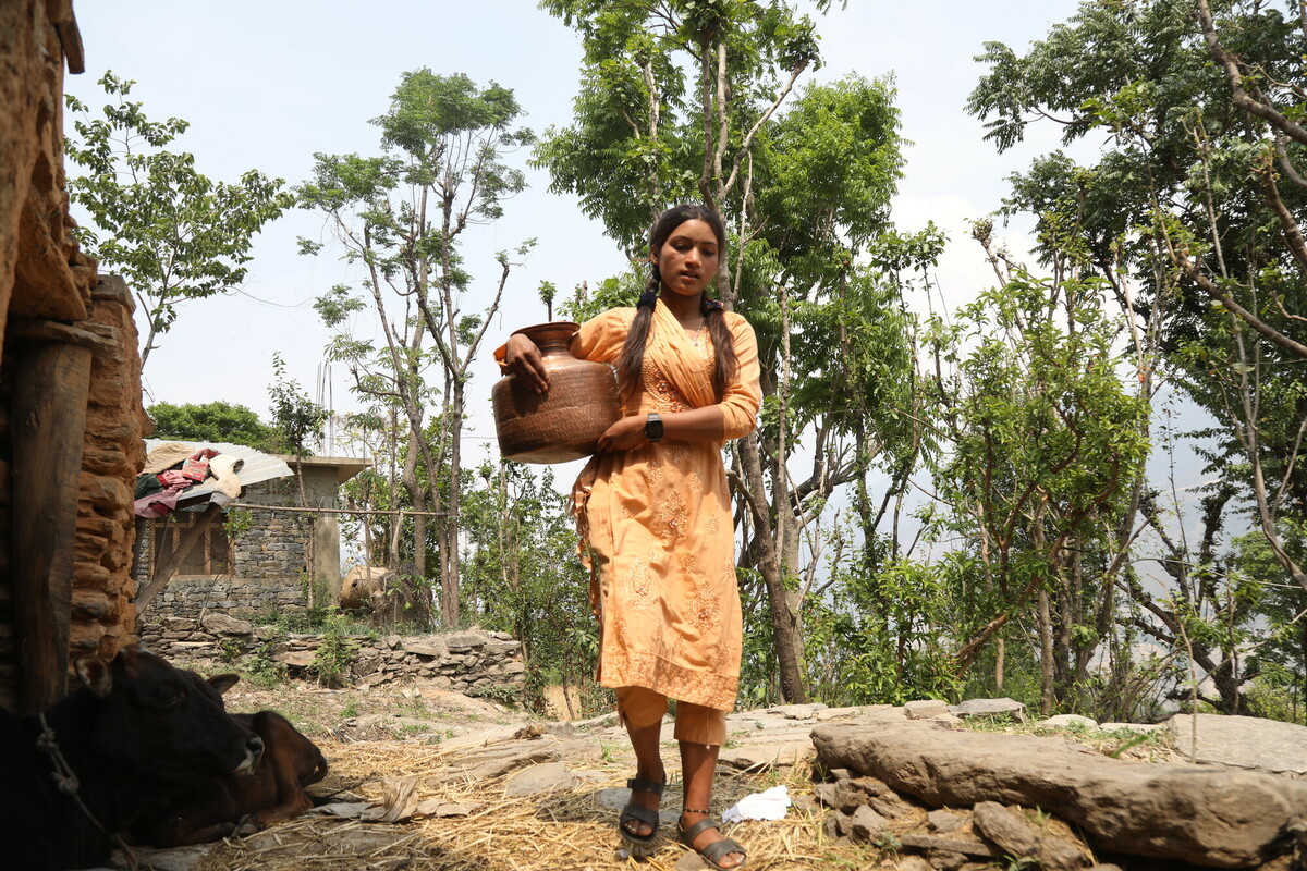 A woman in an orange dress carries a woven basket outdoors, surrounded by trees and a rustic stone structure. She walks on a path with straw underfoot, amid a lush, green landscape.