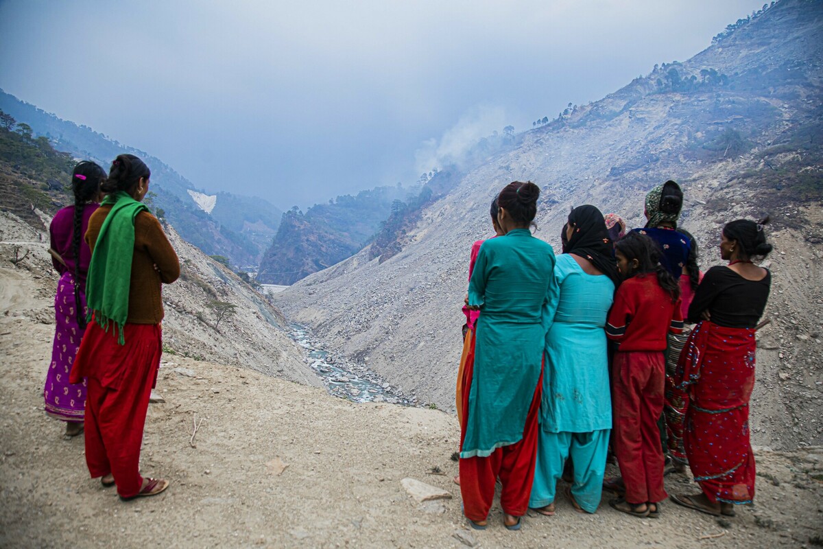 A group of people in colorful clothing stand on a rocky landscape, overlooking a narrow valley with a river. The surrounding hills are covered in dust and smoke, suggesting a recent landslide. The sky is overcast.