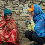 Two women converse outside a small stone building. One is seated, wearing a red shawl and green headscarf. The other crouches, wearing a blue scarf and orange cap. A man in the background uses a phone. Lush greenery surrounds them.