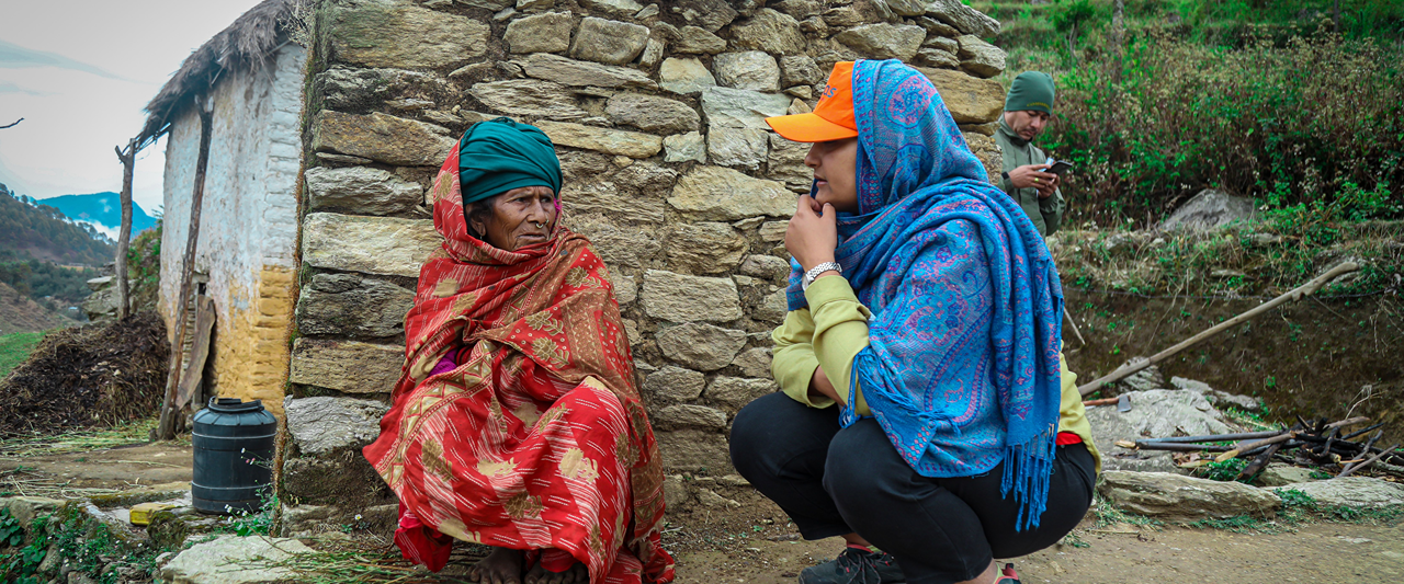 Two women converse outside a small stone building. One is seated, wearing a red shawl and green headscarf. The other crouches, wearing a blue scarf and orange cap. A man in the background uses a phone. Lush greenery surrounds them.