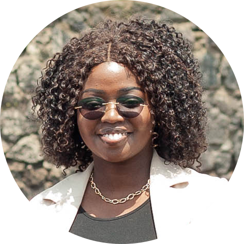 A woman with curly hair and sunglasses smiles against a rocky background. She is wearing a black top, a light-colored blazer, and a gold chain necklace.