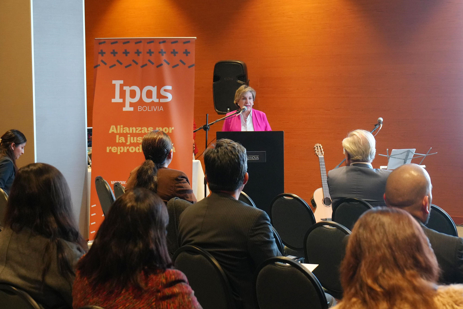 A woman speaks at a podium during an event, with attendees seated and listening. Behind her is a banner that reads "Ipas Bolivia" and "Alianzas por la justicia reproductiva." A guitar rests on a chair nearby.