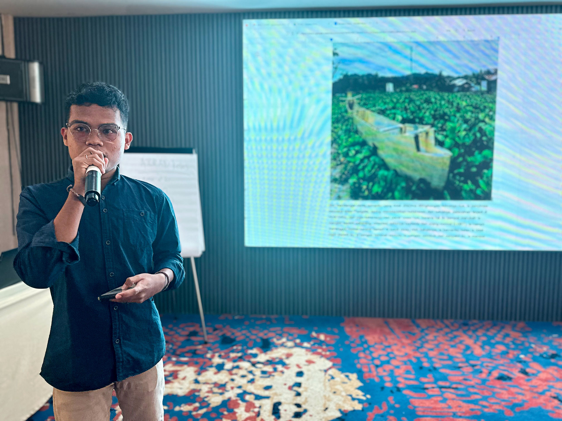 A person holding a microphone stands in front of a screen displaying an image of a lush green agricultural field. A flip chart is visible to the side in the conference room setting.