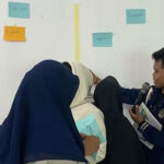 A group of people in dark clothing gathers near a wall filled with colorful paper notes. Aditya is holding a microphone seems to be facilitating or instructing the discussion on topics like Indonesia, child marriage, and school dropout. The notes are labeled "excellent," "great," "good," and "average.