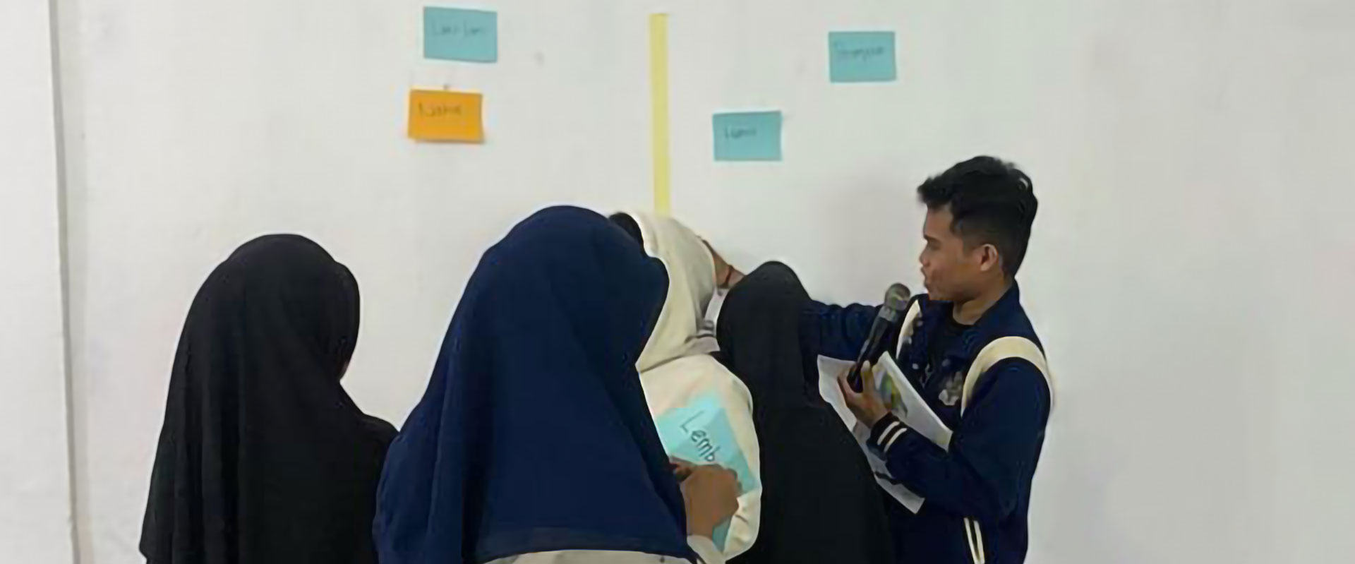 A group of people in dark clothing gathers near a wall filled with colorful paper notes. Aditya is holding a microphone seems to be facilitating or instructing the discussion on topics like Indonesia, child marriage, and school dropout. The notes are labeled "excellent," "great," "good," and "average.