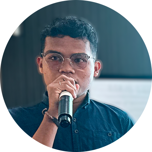 A person with glasses and short curly hair is speaking into a microphone. They are wearing a dark shirt and a bracelet, with a blurred background behind them.