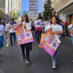 A group of people walking down a city street during a demonstration, holding colorful posters with information. They wear matching white t-shirts and appear focused on their activity. Buildings and graffiti are visible in the background.