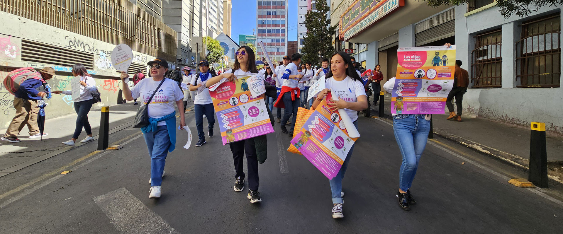 A group of people walking down a city street during a demonstration, holding colorful posters with information. They wear matching white t-shirts and appear focused on their activity. Buildings and graffiti are visible in the background.