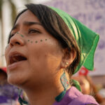 A person wearing a green bandana and earrings shouts during a protest. They have decorative dots on their face. In the background, people hold protest signs with handprints and writing. Palm trees and buildings are visible.