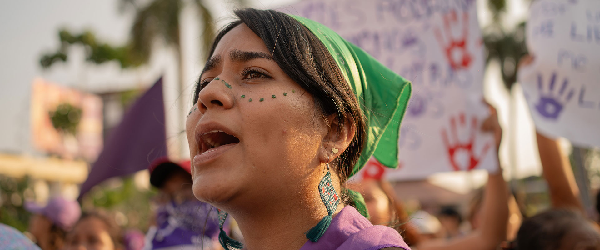 A person wearing a green bandana and earrings shouts during a protest. They have decorative dots on their face. In the background, people hold protest signs with handprints and writing. Palm trees and buildings are visible.