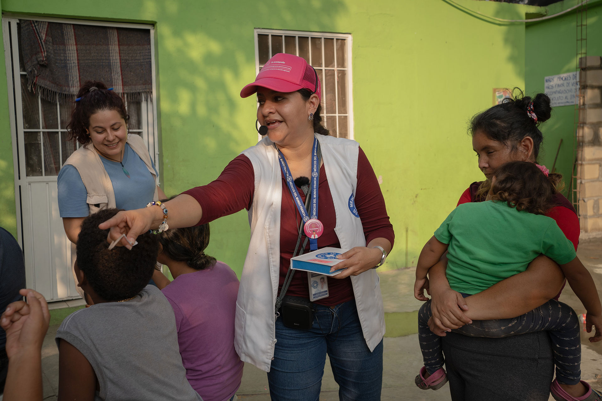 Lourdes Faviel, wearing a pink cap and white vest joyfully interacts with children and adults in an outdoor setting. She holds a booklet and a pen, while a woman nearby smilingly observes and a child is carried by another woman. A vibrant green wall is in the background.