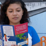 Elizabeth Martínez stands against a colorful mural, reading a pamphlet. The pamphlet features illustrations and text in Spanish, with the words "Te Equivocaste" visible. The person wears a blue and white shirt and has shoulder-length curly hair.