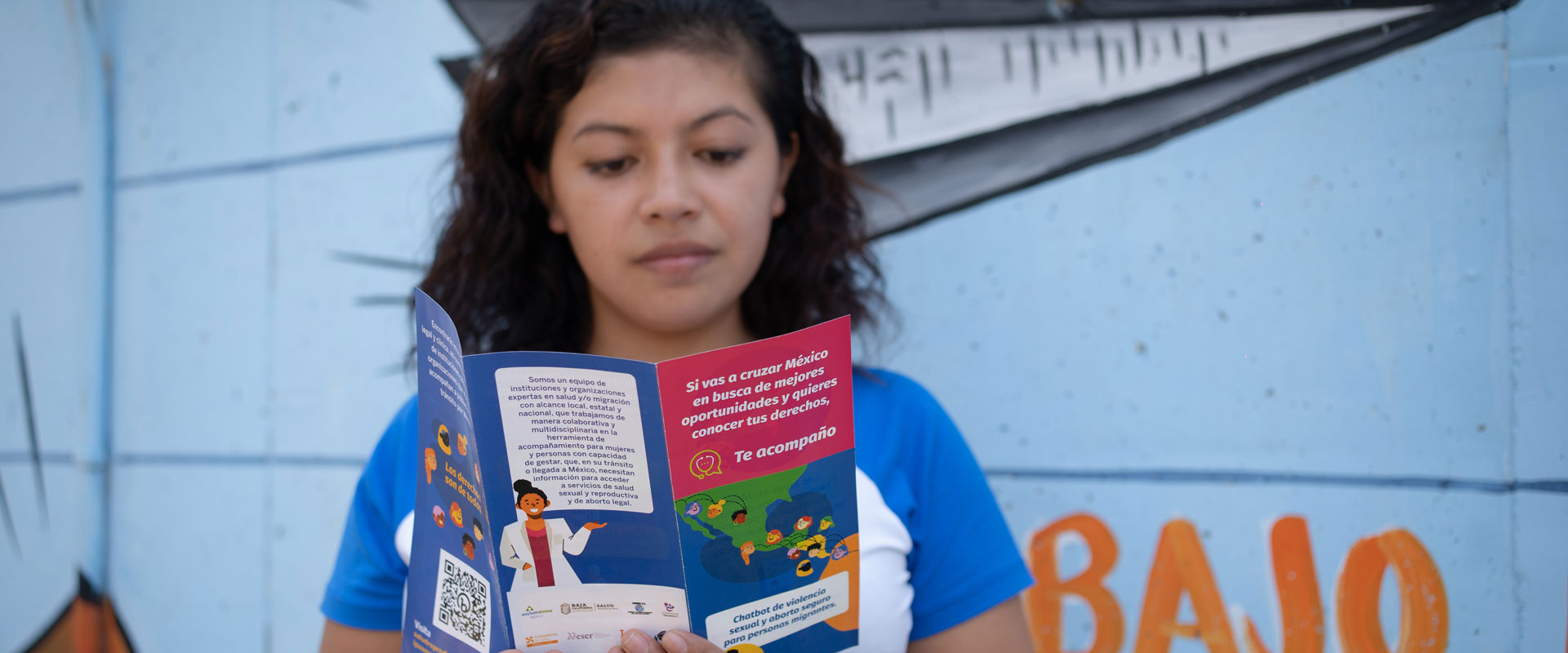 Elizabeth Martínez stands against a colorful mural, reading a pamphlet. The pamphlet features illustrations and text in Spanish, with the words "Te Equivocaste" visible. The person wears a blue and white shirt and has shoulder-length curly hair.