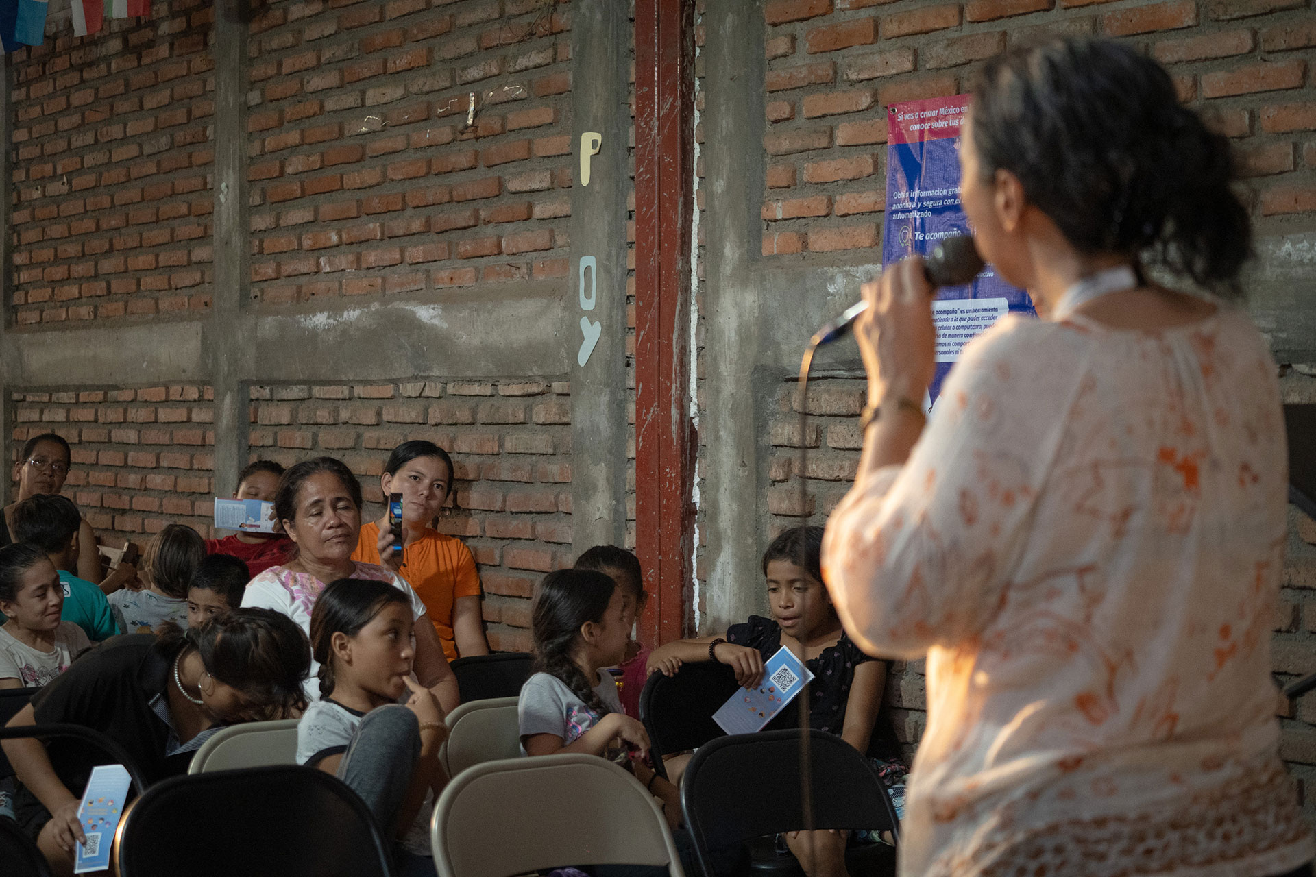 Fabiola Diaz Rovelo  speaks into a microphone in front of a seated audience in a rustic room with brick walls. Adults and children listen attentively, some holding papers. The atmosphere is casual and engaged.