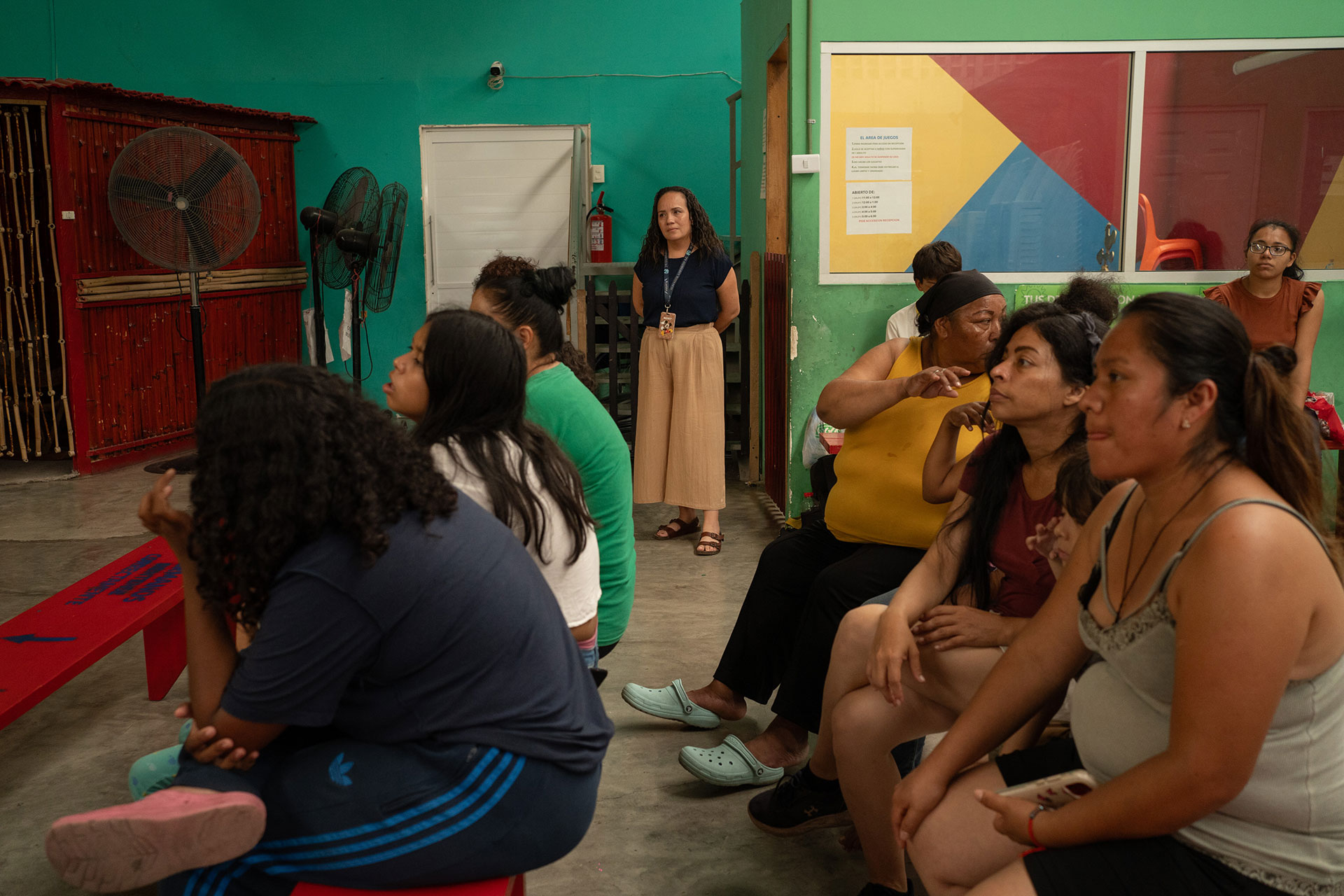 A group of women sits on benches in a colorful room, attentively looking forward. A woman stands in the background, observing the scene. Fans and a fire extinguisher are visible on the wall, along with a partially visible flag.