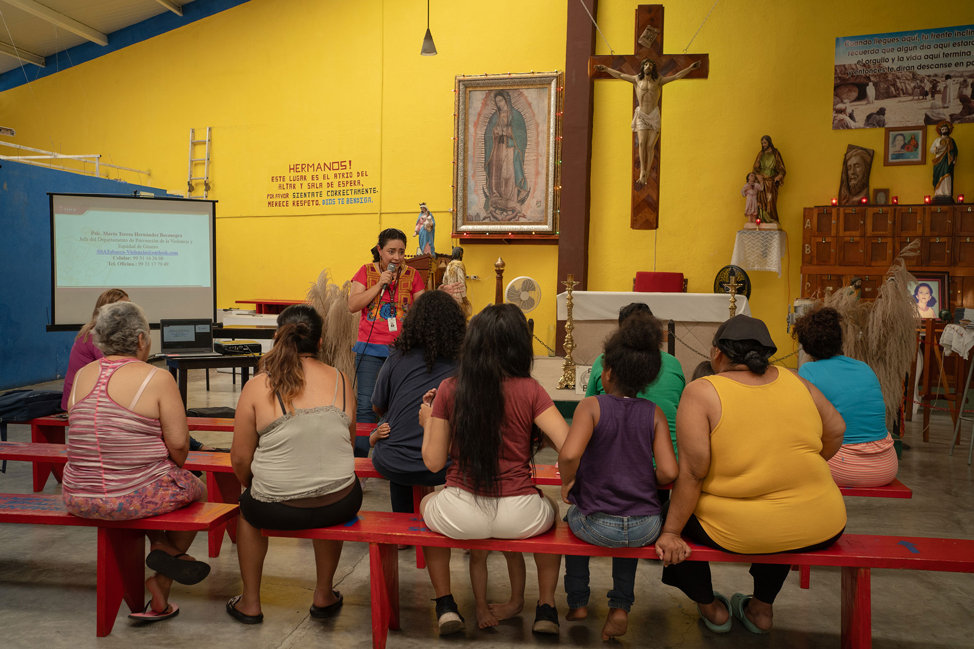 A group of people sits on red benches inside a bright, colorful room with religious decorations. María Teresa Hernández Bocanegra stands at the front leading a discussion. A crucifix and religious paintings are visible on the yellow walls.