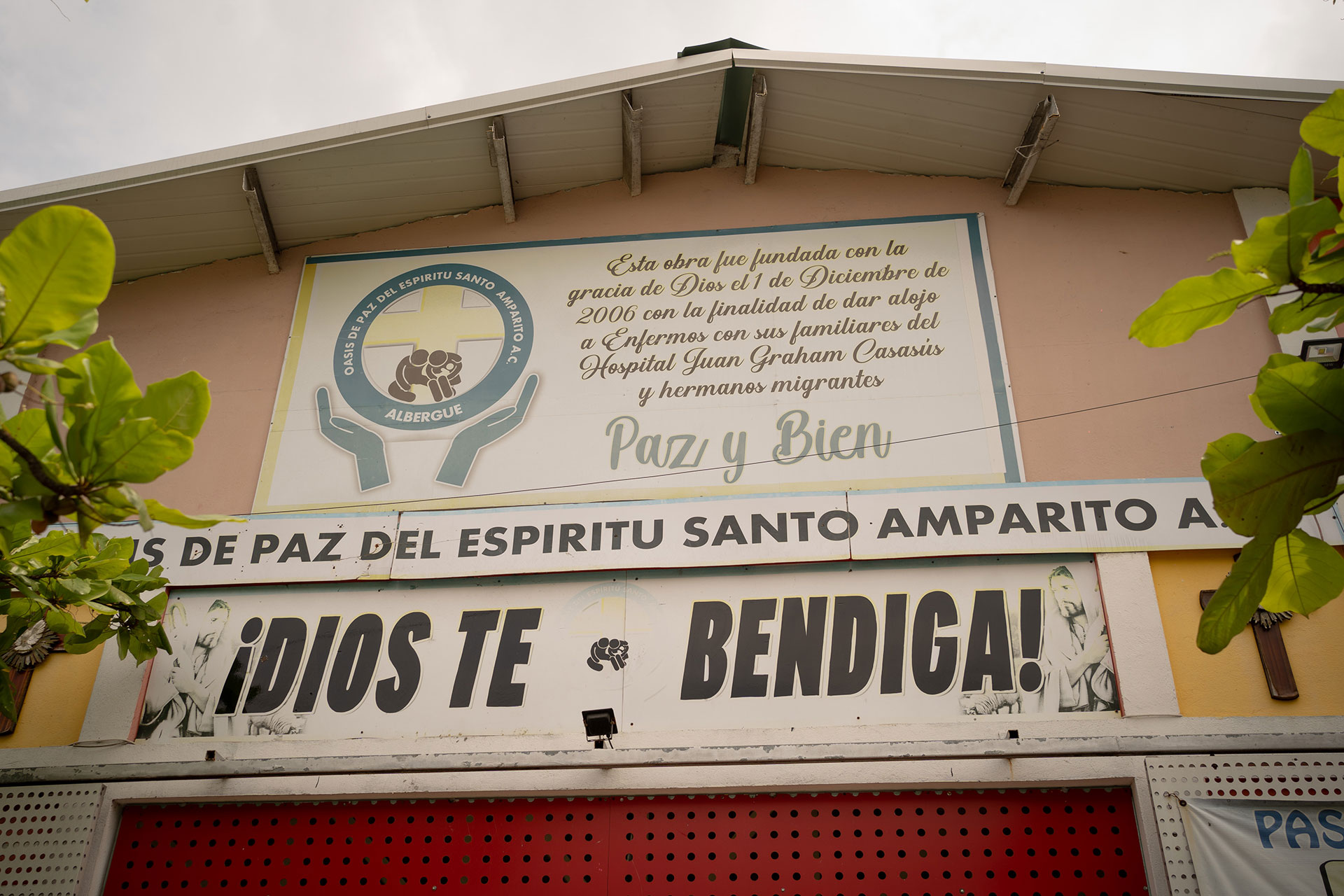 Front view of a building featuring a large sign in Spanish. The building has white walls, a red door, and green foliage in the foreground. The sign mentions "Paz y Bien" and "¡Dios te bendiga!" above the entrance.