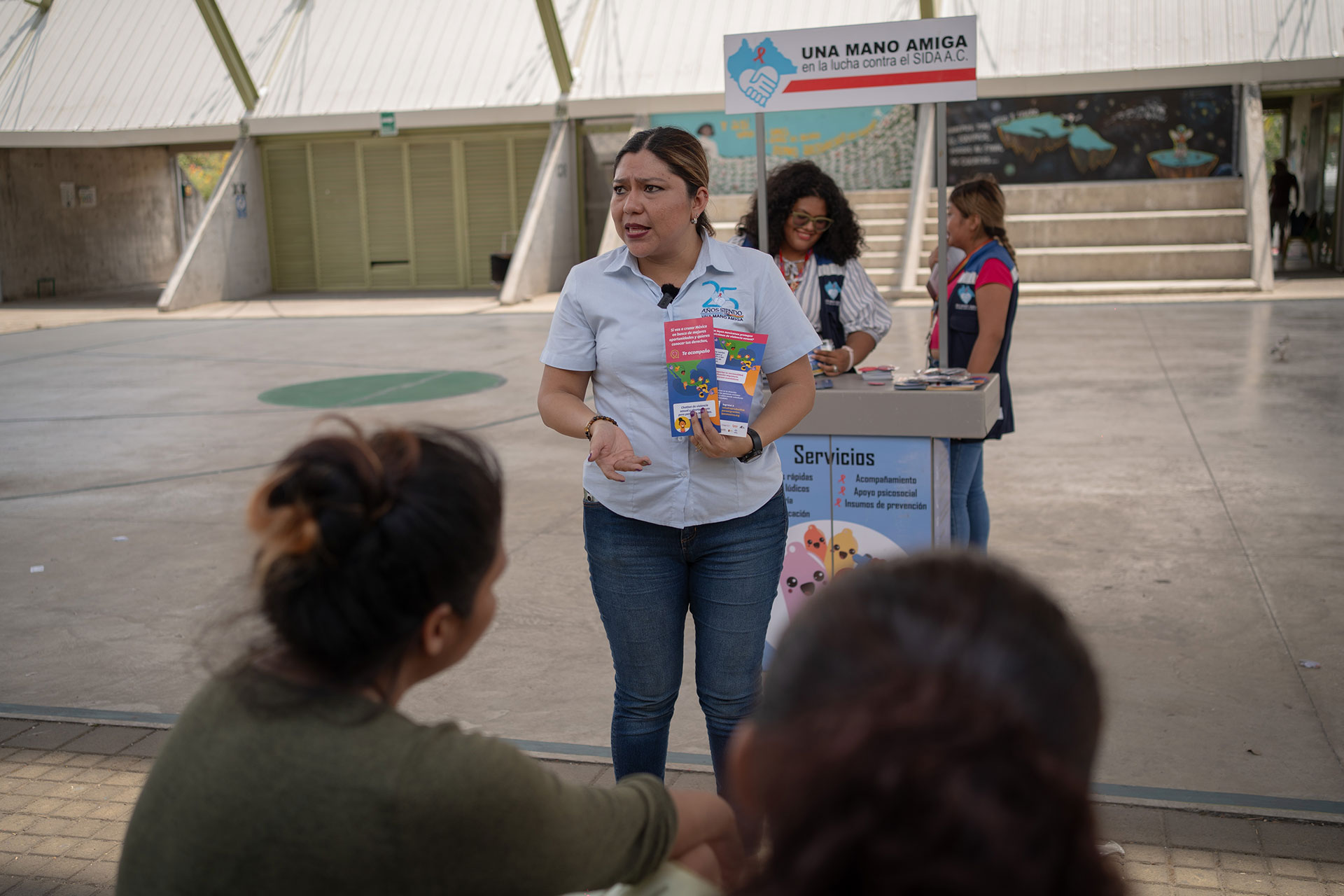 Yadira Guerrero stands in front of a small outdoor information booth, holding colorful pamphlets. Another woman behind the booth is organizing materials. Two people are seated and engaged in the conversation. The setting appears to be a community event.