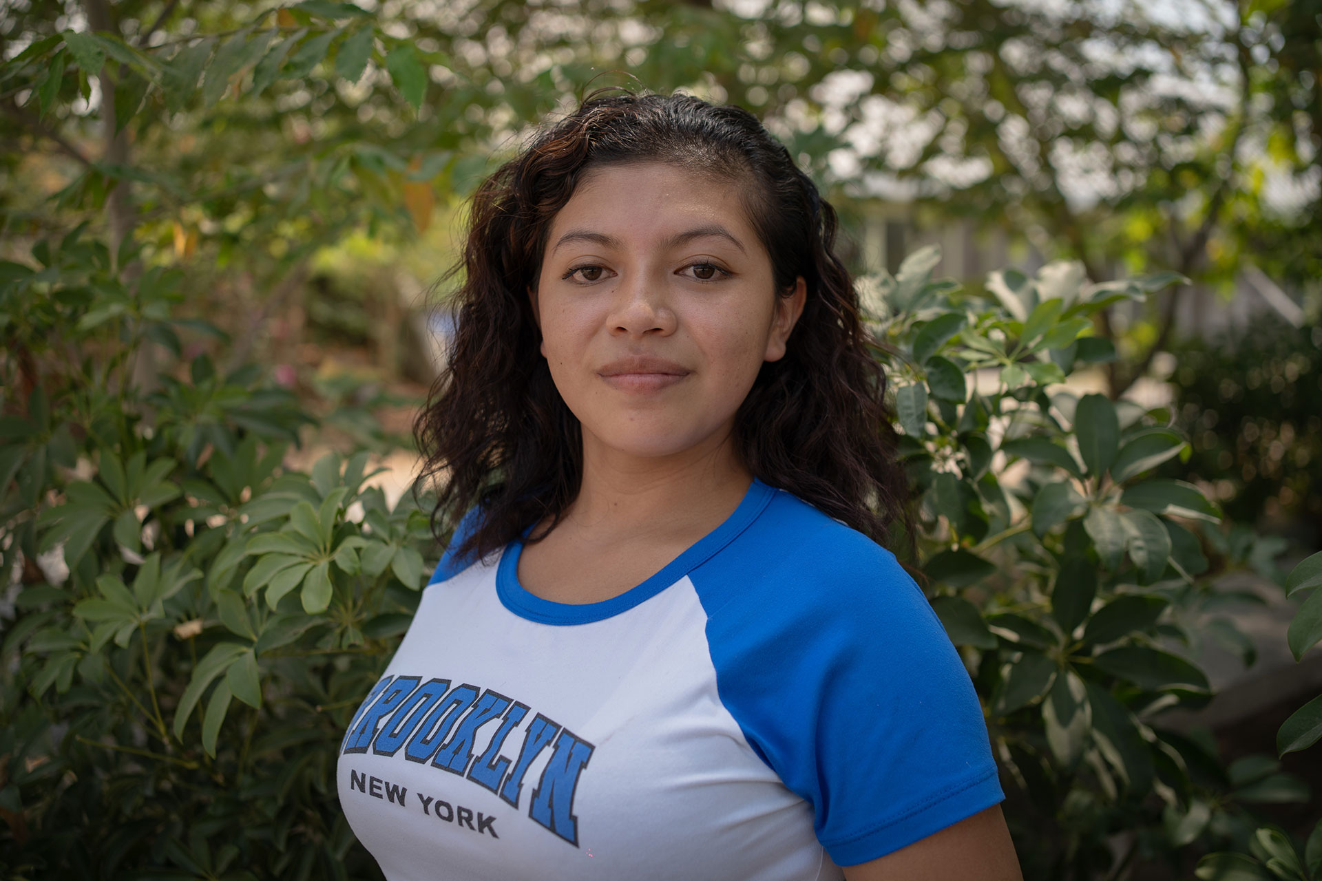 Elizabeth Martínez has long, dark curly hair wearing a blue and white shirt stands in front of green foliage. The shirt has "BROOKLYN NEW YORK" printed on it. The background is softly blurred, highlighting the natural setting.