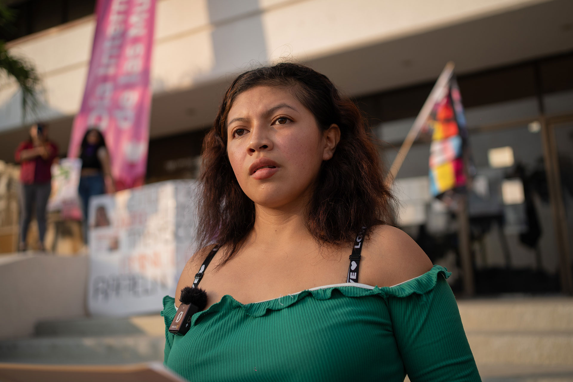 Elizabeth Martínez has long brown hair stands outside, wearing a green off-the-shoulder top. She holds a paper and has a microphone clipped to her shirt. Behind her are blurred buildings, banners, and a multicolored flag.