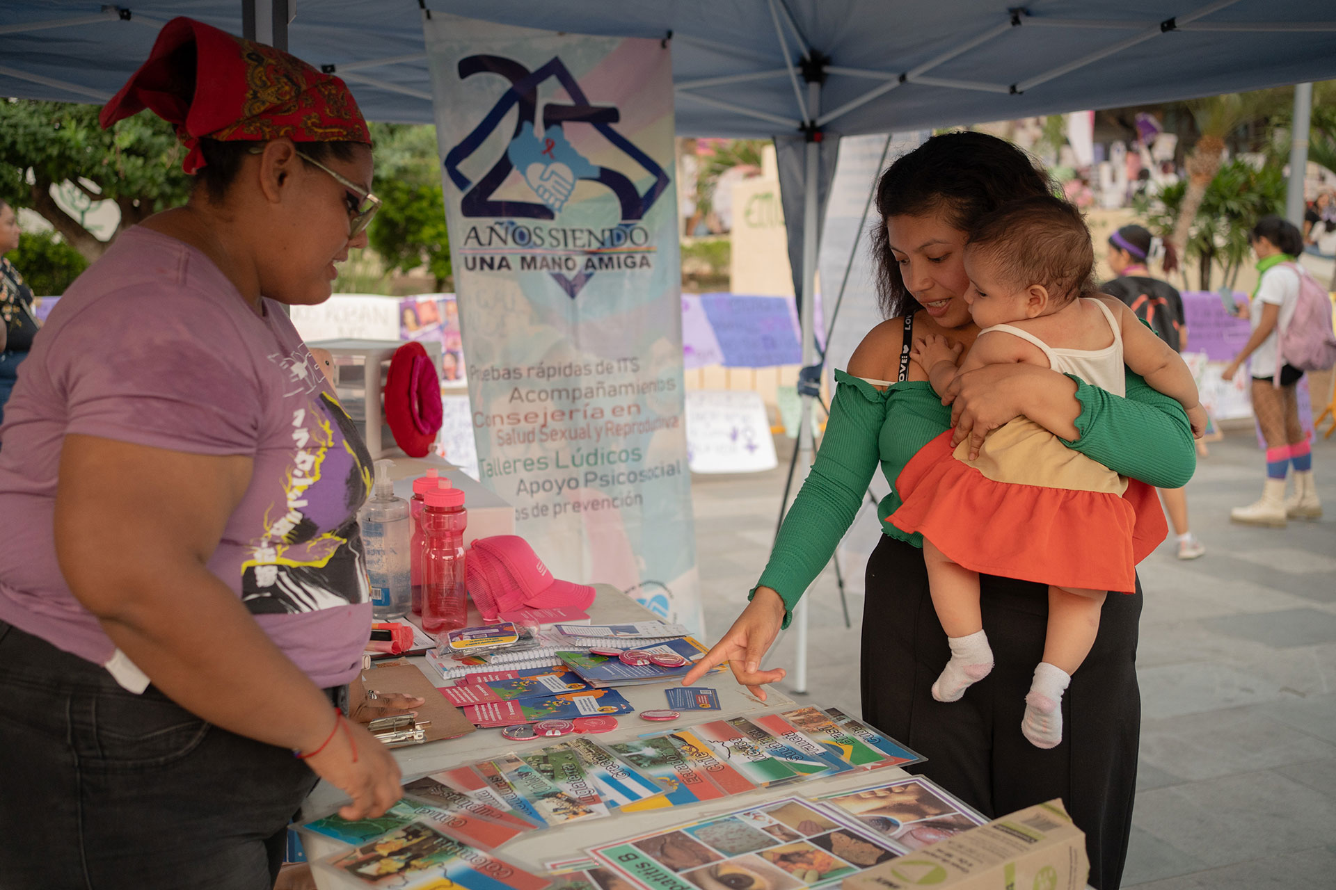 Elizabeth Martínez,  holding a baby talks to another woman at a booth with brochures and a banner under a blue canopy. The setting appears to be an outdoor community event.