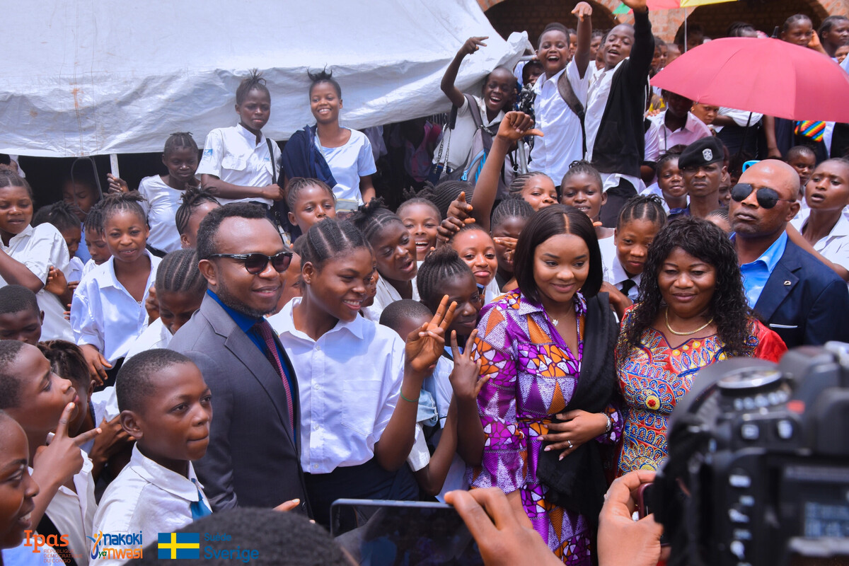 A group of smiling students in school uniforms gather around several adults dressed in colorful attire. The group is outdoors, with one person holding an umbrella. The mood is joyful and celebratory.