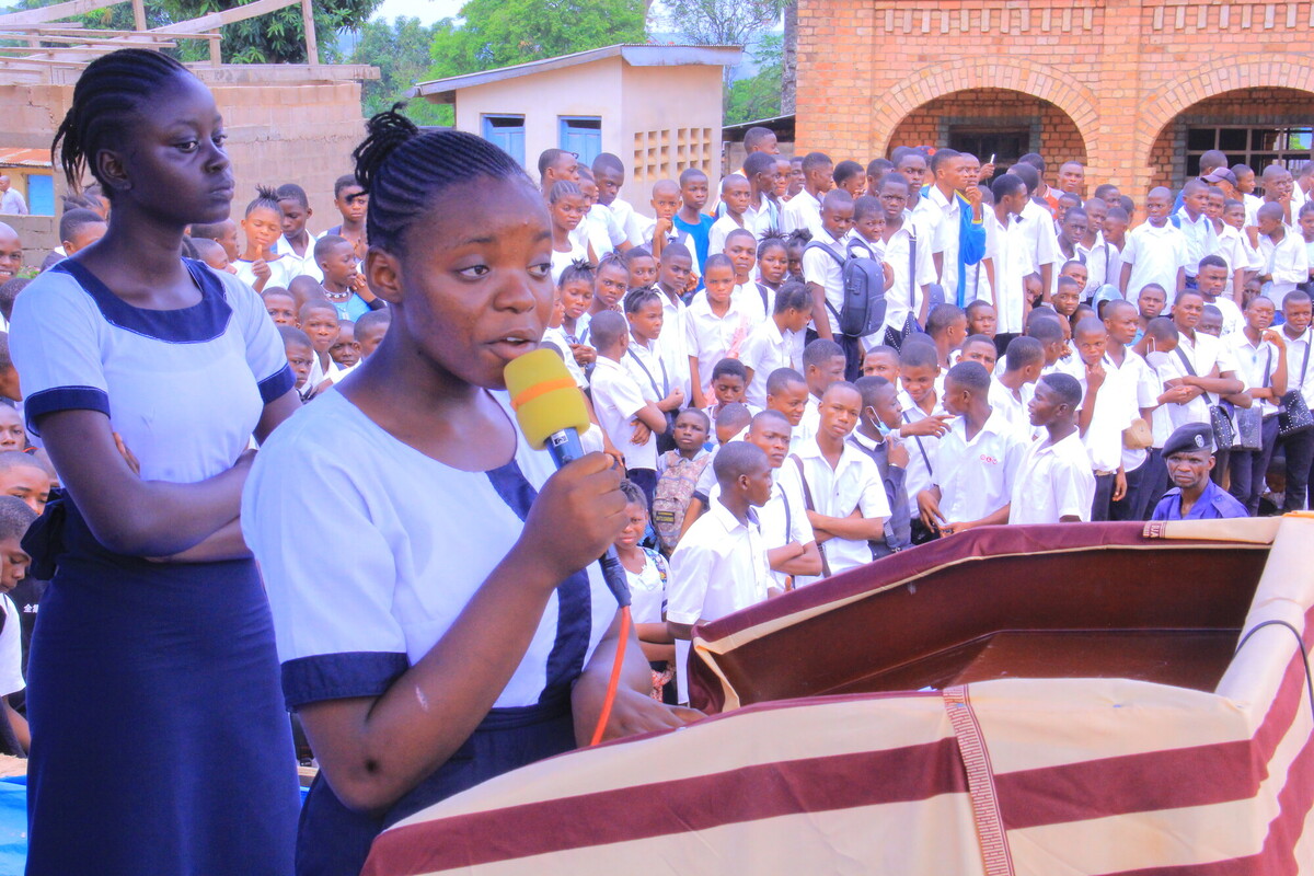 A girl in a school uniform speaks into a microphone at a podium, addressing a large crowd of students in similar uniforms gathered outside a brick building. Another girl stands beside her, listening attentively.