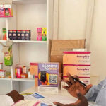 A man and a woman are having a discussion in a pharmacy about medical abortion services in Kenya. The woman, wearing a white coat, stands behind a counter with medicine boxes, including Femiplan. A sign with a QR code is displayed between them.