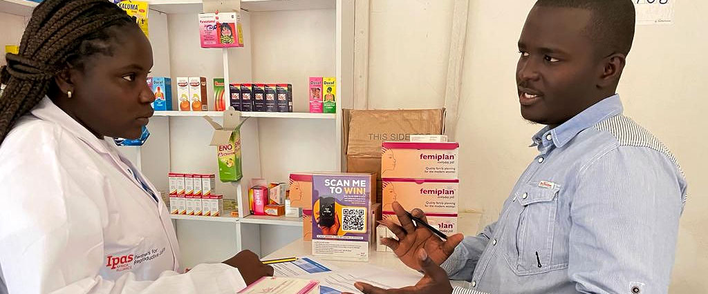 A man and a woman are having a discussion in a pharmacy about medical abortion services in Kenya. The woman, wearing a white coat, stands behind a counter with medicine boxes, including Femiplan. A sign with a QR code is displayed between them.