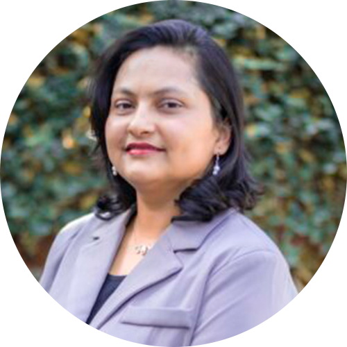 Khusbu Poudel has shoulder-length dark hair, wearing a gray blazer and a black top, is smiling against a blurred outdoor background with greenery.