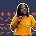 A person wearing a yellow "Community Voices Network" T-shirt speaks into a microphone, gesturing with one hand. The background features a pattern of orange crosses and lines on a dark blue backdrop.