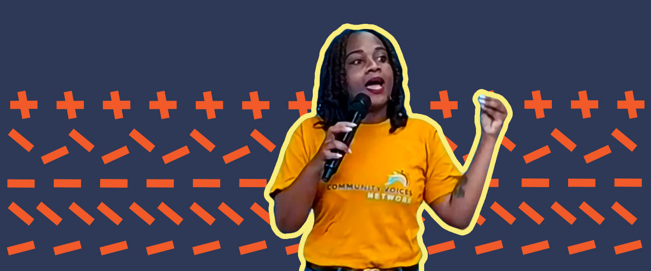 A person wearing a yellow "Community Voices Network" T-shirt speaks into a microphone, gesturing with one hand. The background features a pattern of orange crosses and lines on a dark blue backdrop.