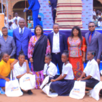 A group of people, including students and adults in formal attire, pose for a photo at an outdoor event. Some hold branded bags. A colorful banner and a crowd in the background suggest a community gathering or celebration.