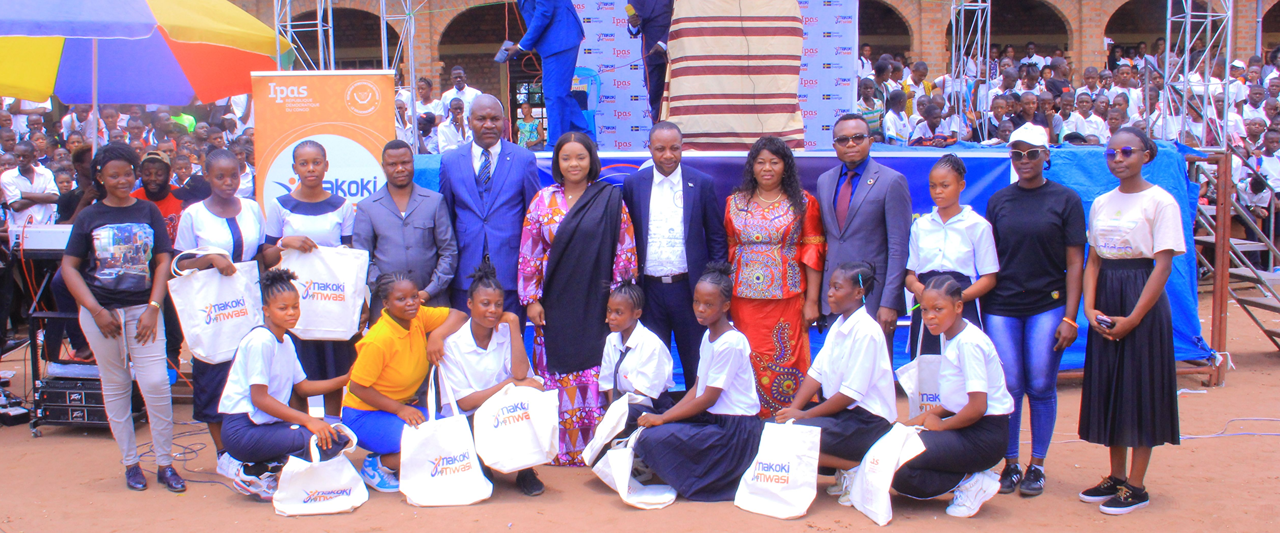 A group of people, including students and adults in formal attire, pose for a photo at an outdoor event. Some hold branded bags. A colorful banner and a crowd in the background suggest a community gathering or celebration.