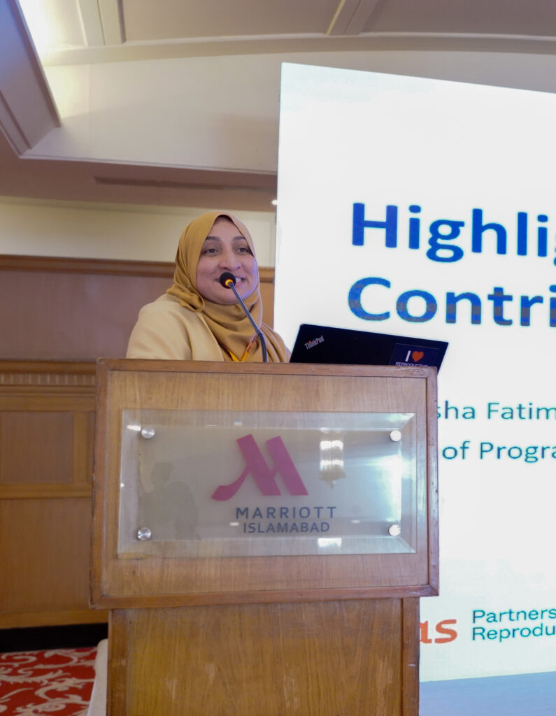 A person wearing a yellow hijab speaks at a podium with a Marriott Islamabad logo. A large screen behind them displays partially visible text in blue.