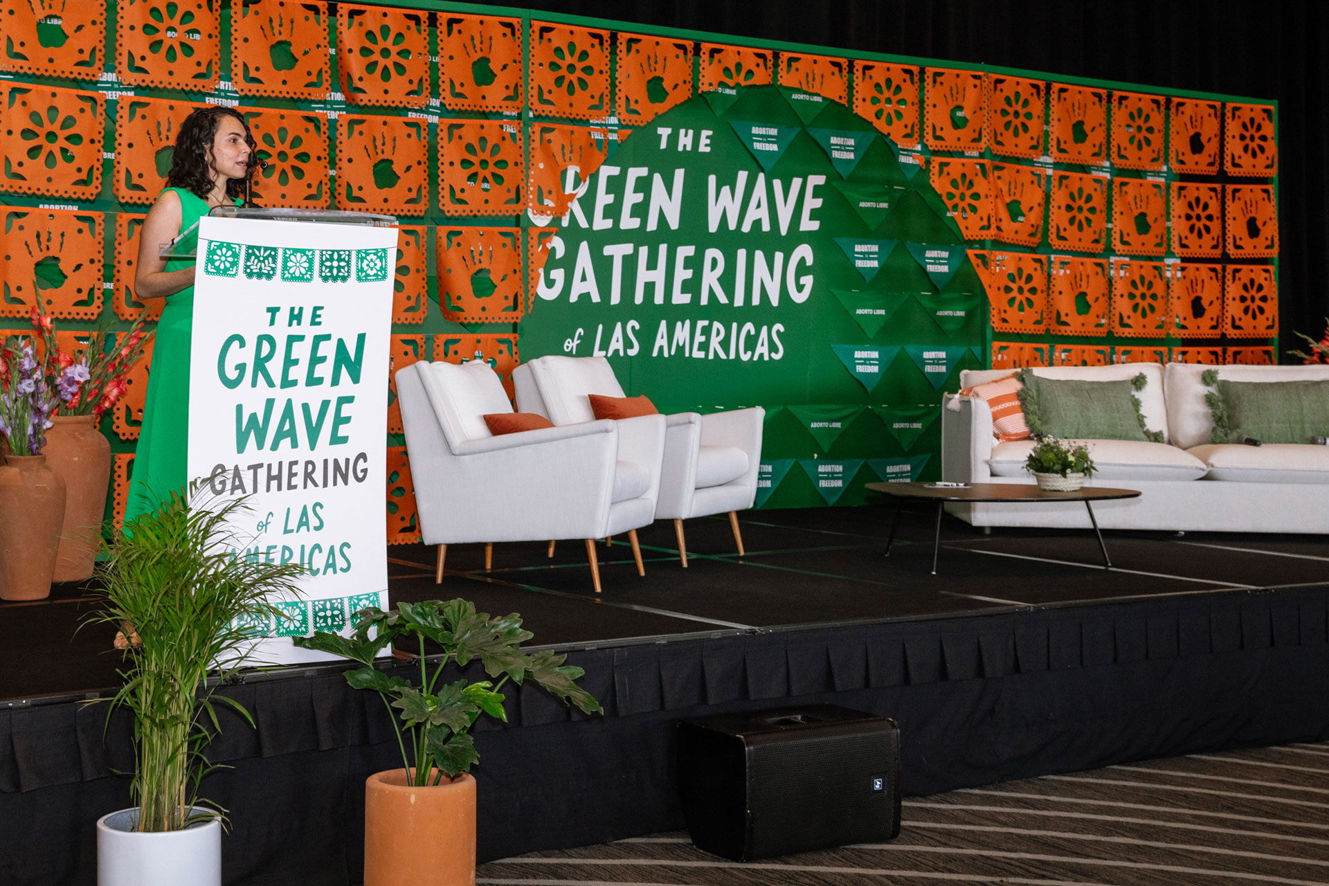 María Antonieta Alcalde Castro, Director, Ipas Latin America and the Caribbean, stands at a podium decorated with "The Green Wave Gathering of Las Americas" text. Beside her are white chairs and a white sofa adorned with green and orange pillows. The backdrop features a vibrant green and orange geometric design, representing the spirit of the Green Wave Gathering.