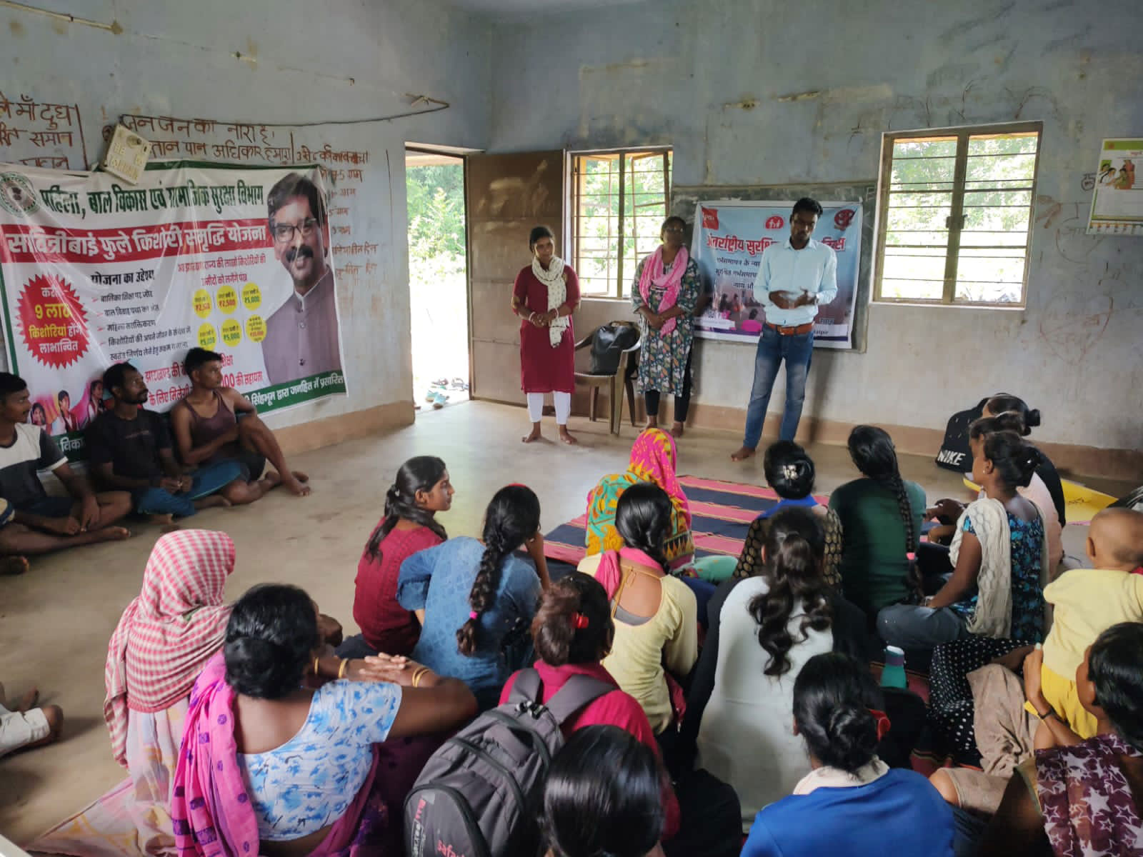 A group of people sit on mats in a classroom, listening to a speaker. Posters with text and images are on the walls. The setting appears to be an educational session or community meeting.