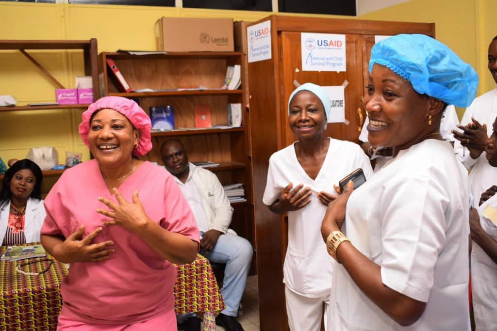 Healthcare workers in a clinic smile and interact. Two women in scrubs and colorful caps engage with colleagues in a room with medical supplies and informative posters on the walls.