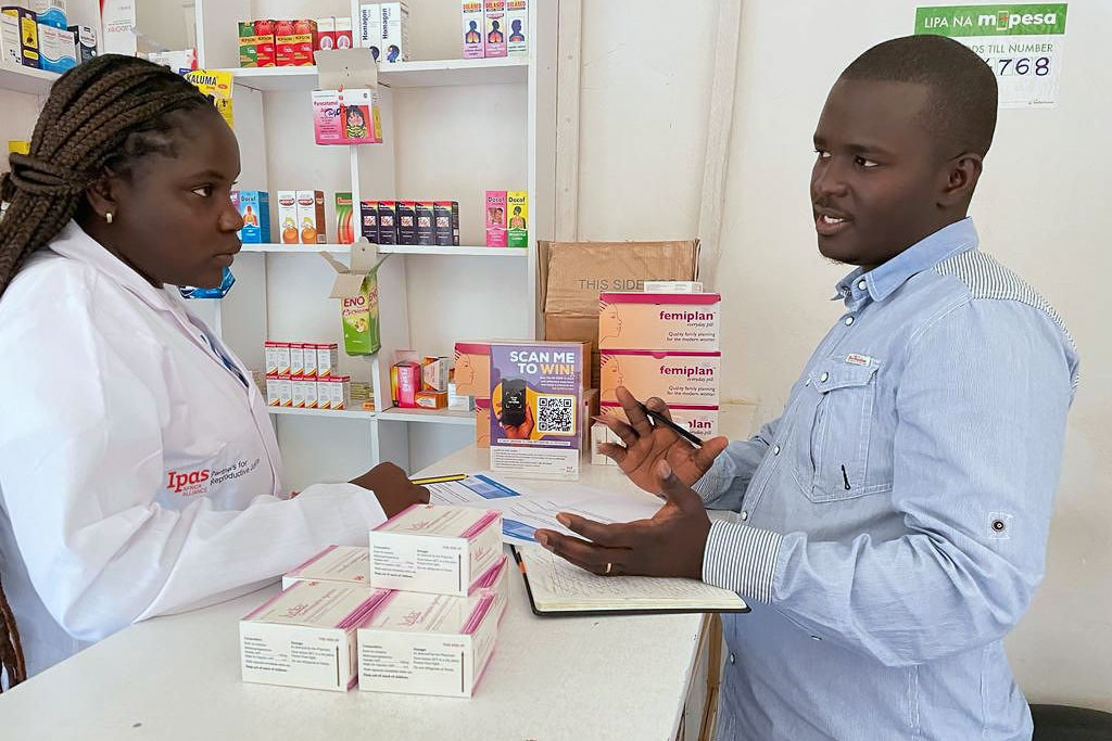 A pharmacist in a white coat discusses medication details with a man holding a pen and notebook. They are surrounded by medicine boxes and health products on shelves in a pharmacy setting.