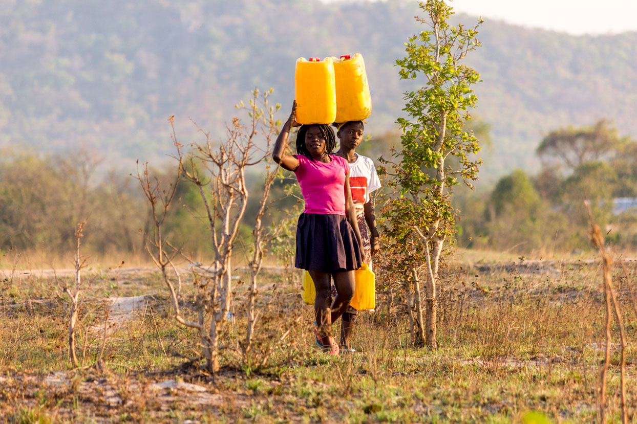 Two people walk through a dry, grassy field carrying yellow jerry cans. One person balances two cans on their head, while the other carries one by hand. Sparse trees and a distant hill are visible in the background.