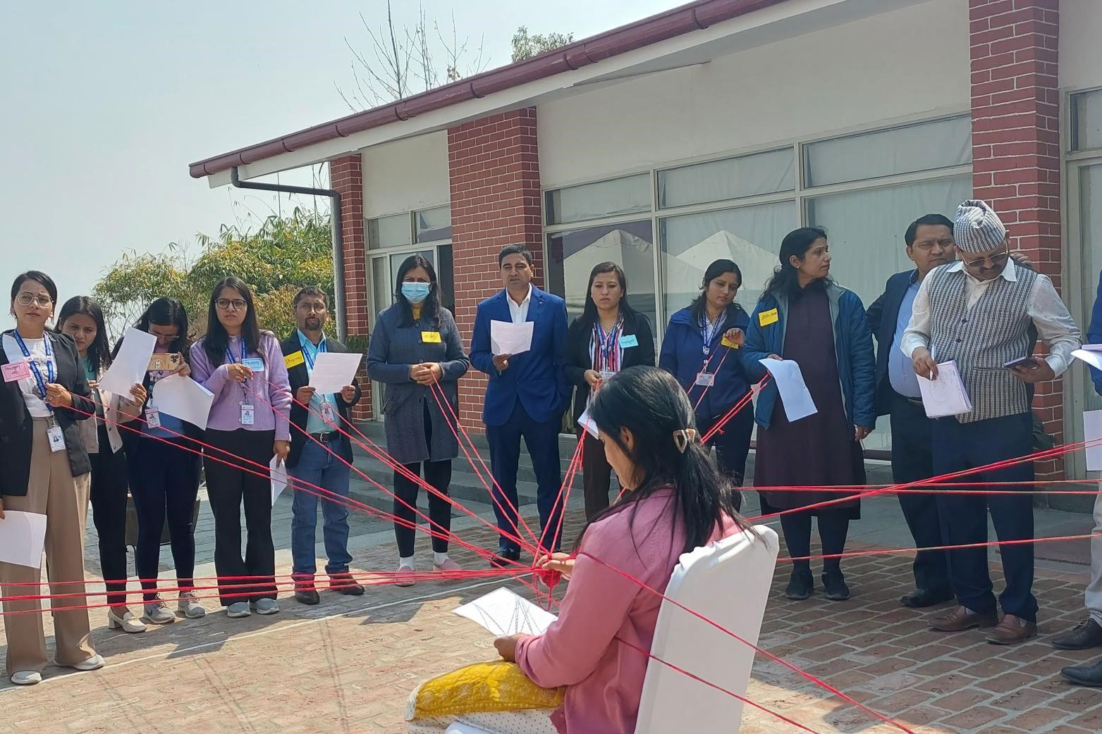 A group of people stand in a circle around a seated individual, each holding red strings connected to the person in the center. They appear concentrated and are holding papers and pens. The activity seems to be taking place outside in front of a building.