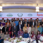 A large group of people, including men and women, pose together indoors in front of a display with the logos of various organizations. The setting includes tables with floral decorations and elegant lighting fixtures.