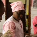 Three women are working in a hospital room. They are wearing pink scrubs and caps. One woman is preparing medical supplies while the others are focused on their tasks. The walls are tiled and there are medical posters in the background.