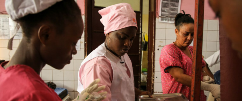 Three women are working in a hospital room. They are wearing pink scrubs and caps. One woman is preparing medical supplies while the others are focused on their tasks. The walls are tiled and there are medical posters in the background.